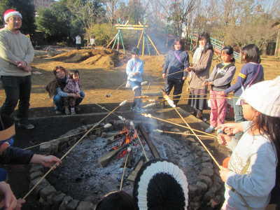 （写真）細長い篠竹の先にパン生地を巻き付けてたき火で焼いている子どもたち.jpg